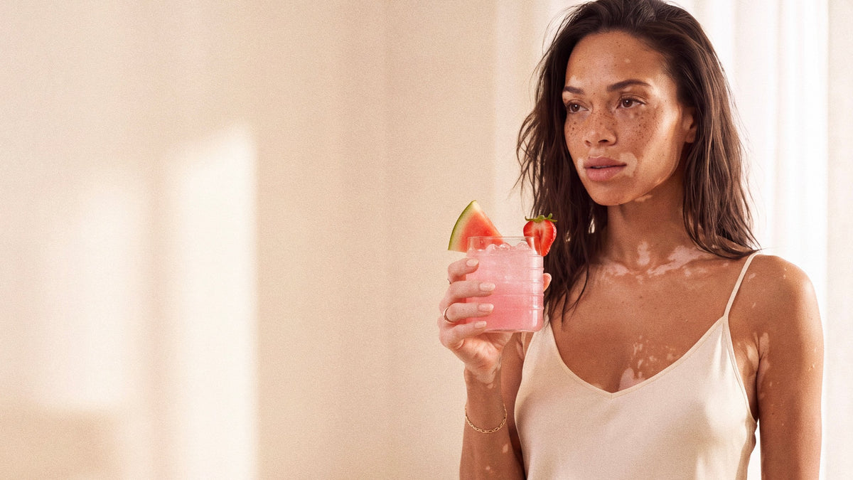Woman in a cream silk camisole holding a glass of pink Lyrenna Strawberry Watermelon drink with a strawberry and watermelon slice