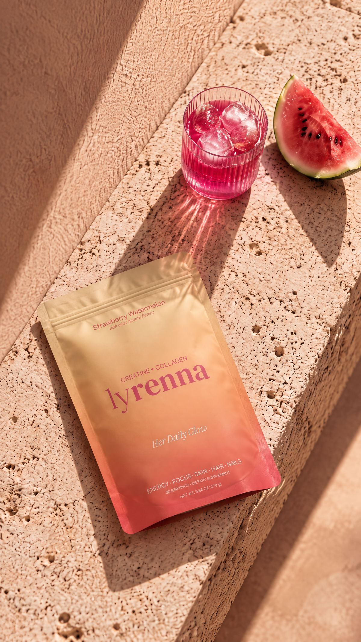 Lyrenna Creatine + Collagen Strawberry Watermelon pouch on a sunlit travertine ledge with a ribbed glass of pink drink and a watermelon slice